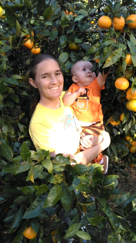 A woman holding a baby stands among orange trees, reaching for ripe oranges in a sunny orchard. The scene captures a joyful moment of family bonding in nature, highlighting the beauty of fruit picking and outdoor activities.